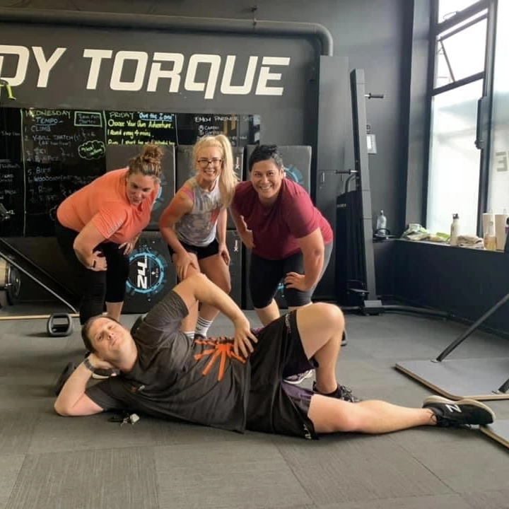 Group of smiling middle age adults at Body Torque Gym posing after a workout, with one man playfully lying on the floor in front of the other members.